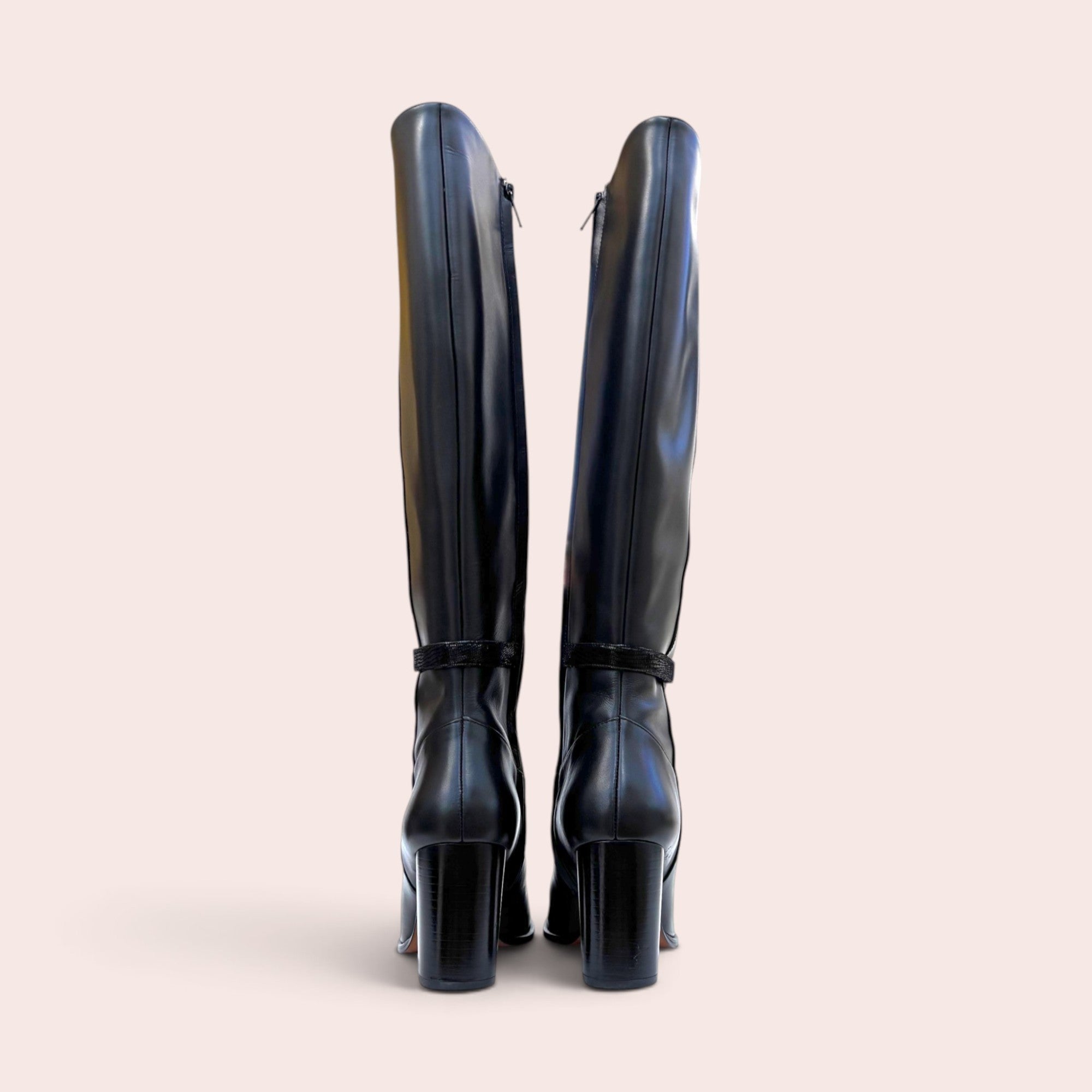 Back view of black leather knee-high boots with block heels and subtle ankle strap detail against a neutral background.